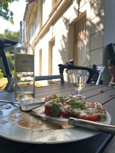 The image depicts a close-up view of a table outdoors, likely on a patio or terrace, with a building in the background. On the table, there is a nearly empty plate with remnants of a meal, a fork, a knife, a glass of white wine, and a partially consumed bottle of white wine. A dish of what appears to be a caprese salad with tomatoes, mozzarella, and basil is also on the plate. The building in the background is light-colored, possibly stucco, with shuttered windows and an overhanging roof. Sunlight casts shadows from trees onto the building. The overall impression is of a leisurely meal being enjoyed outdoors. The location is stated to be in Bièvres, France.