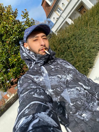 A male individual is depicted in a close-up, upward-angled outdoor photograph taken in Hoenheim, France, under a clear blue sky. The subject, positioned centrally, has a beard and mustache, wears a blue patterned baseball cap, a black and white abstract-patterned puffy winter jacket, and a white in-ear headphone in his left ear. A lit cigarette is held between his lips, extending from the lower right side of his mouth. He gazes directly towards the camera. The background on the left features a tree with dense green foliage, while the right exhibits a tall, manicured green hedge. Above the hedge, a portion of a multi-story residential building with light-colored walls, white-framed windows, and a reddish-brown tiled roof is visible. The scene is brightly illuminated by natural sunlight, indicating a clear day.