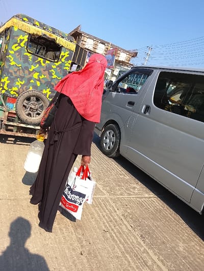 The image depicts a street scene in Shibpur, Bangladesh, under a clear, bright sky. A person, viewed from behind, is walking on a paved surface, moving from left to right across the frame. This individual is dressed in a full-length black abaya, secured with a visible tie-belt, and a vibrant red hijab covering their head and shoulders. They are carrying a transparent plastic bottle containing a white liquid in their left hand and two white shopping bags with red handles in their right hand. One bag displays the word "Chittagong" and "Free Gallery."
To the immediate left of the person, a three-wheeled auto-rickshaw or similar utility vehicle is stationary. Its body is covered in a camouflage pattern of greens, browns, and black, with a yellow fringed canopy bearing the word "APEX." A spare tire is mounted on the rear. Further to the right, a silver-grey minivan, possibly a Toyota Hiace, is parked or moving slowly. Its passenger side sliding door appears partially ajar or recently closed.
In the background, multiple utility poles with overhead wires are visible against the blue sky. Distant buildings, appearing to be multi-story structures with some exposed brick or concrete, line the street. The ground is a light-colored concrete or asphalt, showing texture and some faint lines, suggesting a roadway or wide pedestrian area. A distinct shadow of a person, likely the photographer, is cast on the ground in the lower-left foreground. The overall lighting suggests midday.