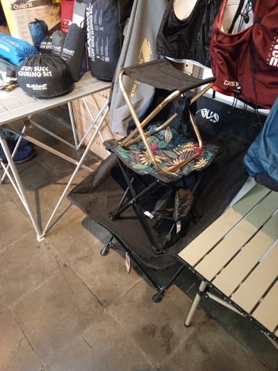 This image depicts an indoor retail display of outdoor and camping gear in Baza, Myanmar (Burma). The foreground features a floor composed of large, irregular gray concrete or stone tiles. In the midground, on the left, a light-colored folding camping table with a slatted top displays several black stuff sacks, one prominently labeled "STUFF SUCK COOKING SET" by "SEATO SUMMIT". To the right of this table, a stack of folding camping chairs or stools is visible: a black fabric stool with a gold-colored aluminum frame, partially folded, rests on top of a similar folding stool featuring a tropical leaf pattern (green, pink, and yellow foliage on a dark background) and a gold-colored aluminum frame. Both stools are positioned on a larger black folding camping cot or chair. A light tan/beige folding camping table with a slatted top is partially visible on the far right. The background exhibits various camping equipment displayed against a wall, including rolled sleeping bags, backpacks, and other accessories. Visible text on these items includes "SLA SUMMER OUTDOOR GEAR" and "FLYWEIGHT POLE". A white box with the word "avenil" is partially visible in the upper left background. No individuals are present in the scene.