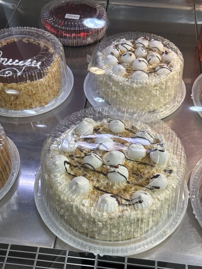The image shows a display of cakes in a store. In the foreground, a large round cake is covered in white frosting and decorated with meringue dollops drizzled with chocolate. Behind it and to the left, another round cake with chocolate frosting and chopped nuts is visible. In the background, a round cake with a red topping is in a clear container, and a partially visible cake with white frosting is to the right. The display appears to be in a refrigerated case with a metallic surface. The context of Vladikavkaz, Russia is not discernible from the image itself.