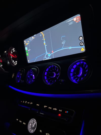 The image shows the interior of a modern car at night, with a focus on the dashboard and infotainment screen. The infotainment screen displays a navigation map, indicating the car is in or near Krumovo, Bulgaria, with a speed limit of 75 km/h highlighted. The map shows roads, buildings, and points of interest like "Airsoft Plovdiv". The car's interior is illuminated by ambient blue lighting, particularly around the air vents, which are glowing with a purple-blue light. On the dashboard, there are illuminated circular gauges with red needles, the climate control panel with illuminated buttons and icons, and a central clock with Roman numerals. The overall scene is dark, with the primary light sources being the car's internal lighting and the infotainment screen. No people are visible in the image.