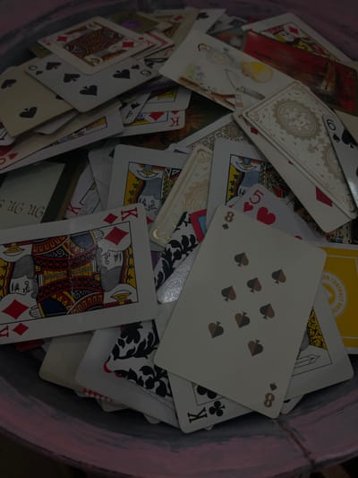 A chaotic pile of playing cards fills the frame, seemingly scattered at random. Numerous card faces are visible, including an 8 of spades, a 5 of hearts, a King of diamonds, and a King of clubs. Other discernible cards feature illustrations and ornate designs, along with numerical and alphabetical markings such as "8", "K", "A", "5", and "6". The phrase "LAN CONTRACT BRIDGE" appears on one card. The cards are stacked and jumbled together in a circular container.