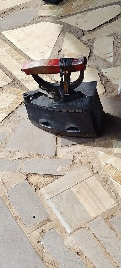 An antique, black, cast iron clothes iron with a red wooden handle is situated on a patterned ground made of irregular, flat stones. The iron is positioned on its side, revealing two rectangular openings on its body. The handle is secured by dark, wrapped material. The ground beneath the iron is a mosaic of light brown, beige, and grey stones of varying shapes and sizes, with visible dirt and debris in the gaps between them. The lighting suggests an outdoor setting, possibly a courtyard or walkway. The overall scene appears static, with no visible activity. The location is specified as Kachia, Nigeria.