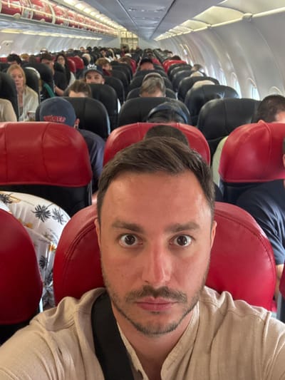 A medium, candid, and slightly distorted eye-level shot shows a man on a packed airplane, likely during boarding or disembarking. The man, who takes up most of the right half of the frame, has short, dark hair, light skin, and a goatee. He is wearing a light tan shirt and has a black strap, possibly from a bag, over his shoulder. His eyes are wide open, as if looking directly at the viewer.
The plane interior has red headrests, and the rows of seats are filled with passengers. The people have dark and light hair, and their faces are turned downwards. The photo may have been taken on a flight to or from Labuan Bajo, Indonesia. The airplane aisle is visible on the left side of the frame.