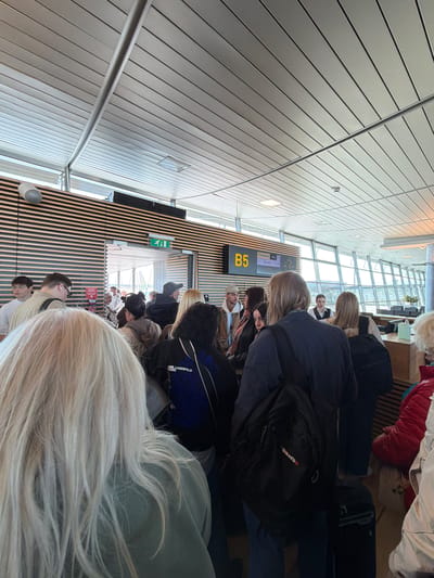 The image captures a crowded airport gate waiting area, likely in Rīga, Latvia. The perspective is from within the throng of people, looking forward towards the gate and large windows.
**Subjects:** Numerous travelers are present, predominantly seen from behind or in partial views. They are clad in various casual attire. Many are carrying personal luggage, including backpacks and other bags. The environment consists of modern airport architecture, featuring light-colored, ridged ceiling panels, vertical slatted wooden walls, and large floor-to-ceiling windows that provide natural light. A metal railing stretches horizontally across part of the scene.
**Activity or Event:** The primary activity is people waiting, presumably for a flight. Some individuals appear to be standing in a queue near the gate, while others are seated on benches. The general atmosphere suggests anticipation or routine waiting.
**Notable Details:** The scene is brightly illuminated by natural daylight entering through the large windows, indicating it is daytime. The contemporary design of the terminal suggests a modern facility. Emotions are not distinctly visible due to the perspective, but the crowd appears orderly.
**Visible Text:** A prominent yellow sign displays the gate number "B5". Above it, a digital departure board is visible, showing "RYANAIR" as an airline, along with labels like "ORIGIN" and "DESTINATION," although specific flight details are illegible. A digital clock is also present on the board. On a black bag or jacket, the text "KARL LAGERFELD PARIS" is discernible, and another black backpack features the word "OMATSU." A standard green exit sign with a running man symbol is mounted on a wooden wall. A red sign with a fire extinguisher symbol and some unreadable text is also visible on the wall.
