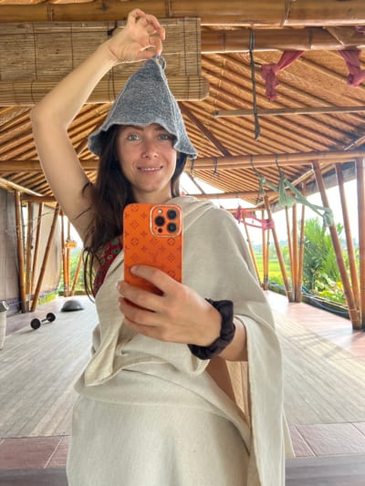 A young woman is taking a selfie with a phone in a natural, open-air structure in Singakerta, Indonesia. The woman is smiling, holding up a gray knit hat. She is wearing a light-colored linen garment. Her phone has an orange patterned case. The structure appears to be made of bamboo and thatch. The background shows a view of rice paddies and greenery. The area seems to be a resort or retreat. There are weights visible in the background, hinting at a fitness area.