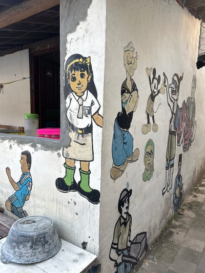 In Banguntapan, Indonesia, a mural is painted on the side of a concrete building. The mural depicts various cartoon characters and people. On the left side, a young girl in a school uniform with a yellow ribbon in her hair is painted. Next to her, a young boy in a blue soccer jersey is kneeling. On the right side, the mural features several characters including Popeye, Mickey Mouse, and a woman wearing a hijab. Below these characters, a person in a construction worker's attire is shown. The building appears to be in a semi-developed area with a sidewalk visible in the foreground. Various objects, such as a large metal basin and plastic containers, are also present in the scene.