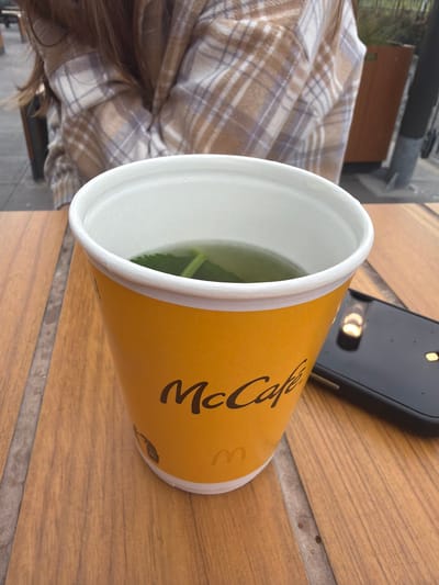 A McCafe cup with a beverage sits on a wooden table. A black smartphone is to the right of the cup. In the background, a person wearing a plaid shirt is visible. The setting appears to be an outdoor cafe at Schiphol, Netherlands.