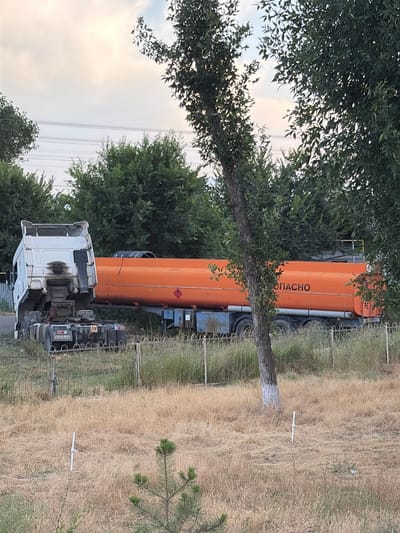 This media file depicts a white truck with an orange tanker trailer parked in a grassy, rural setting. The tanker is labeled with the Cyrillic text "СПАСНО," which translates to "SAVED" or "RESCUE." The truck appears to be a commercial vehicle, possibly a fuel or chemical transporter, given the nature of the tanker. The scene is set outdoors, with trees and dry grass surrounding the vehicles. A wire fence runs along the right side of the frame, and power lines are visible in the distance against a cloudy sky, suggesting it is daytime. The overall impression is one of a functional, if somewhat isolated, location. There are no people visible in the image. The grass appears dry and somewhat unkempt, hinting at either a dry season or a less maintained area. The image is oriented vertically, with the truck and tanker appearing to be on a slight incline or the camera tilted.