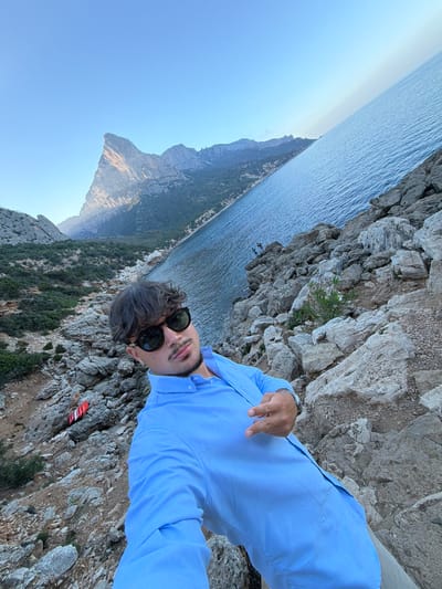 A man wearing sunglasses and a light blue collared shirt is taking a selfie on a rocky coastline with the sea on his right. He has dark hair and a mustache. The background shows a rugged landscape with mountains and dense vegetation. The sky is clear and blue. A striped red and white object is visible on the rocks to his left. In the distance, there are a few people standing on the rocks near the water. The image appears to be taken during daylight. The location is described as the coast of Baunei, Italy.