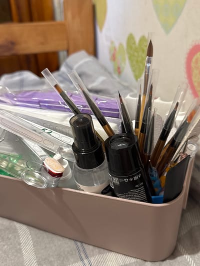 The image depicts a close-up view of a light pink organizer box filled with various nail art supplies. The organizer contains an assortment of brushes with clear protective sleeves, bottles of nail polish or gels, and other small tools. A black bottle with Russian text is prominently visible in the center. The background shows a wooden chair and a patterned cloth or curtain with heart designs. The overall impression is of an organized collection of items for nail art, likely in a personal space. The context of Яромінскі сельскі Савет, Belarus, is not visually discernible from the image itself.