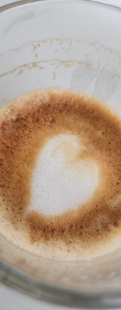 A close-up shot focuses on a heart-shaped latte art design in a clear glass cup, with hints of winged designs on the cup's interior. The coffee foam displays a gradient of light brown to dark brown, with a lighter, foamy center forming the heart. The background shows a white surface with subtle golden specks and the faint impression of angel wings etched into the glass. The scene is indoors, possibly in a cafe or home setting in Fort Myers, United States of America.