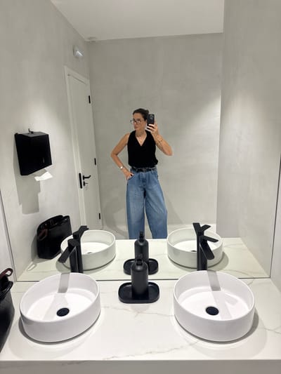 The image depicts a modern, minimalist bathroom with two white vessel sinks on a marble countertop. A woman, wearing a black sleeveless top, denim wide-leg pants, and glasses, is taking a mirror selfie with her smartphone. She has her left hand on her hip and her right arm extended, holding the phone. Next to the sinks are black soap dispensers. On the wall, to the left, is a black dispenser, possibly for paper towels, with a roll of white paper extending from it. A black handbag is placed on the countertop to the left, and another is visible on the floor. A white door with black hardware is positioned on the left side of the room, and a light fixture is mounted on the wall above it. The bathroom walls have a textured, grey finish. The scene is reflected in a large mirror that spans the width of the countertop. This image was taken in a public restroom in Torres Vedras, Portugal.