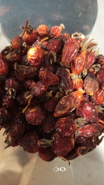 The image shows a close-up view of dried rosehips in a transparent container. The rosehips vary in color from reddish-brown to dark red. Some have stems and remnants of the flower calyx attached. The background is out of focus. There is no evidence to place the image in Юдино, Russia.