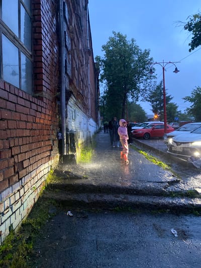 A young child in a pink hat and pink outfit is standing on a wet sidewalk, appearing to be playing in a spray of water. The child is in motion, leaning back with their arms outstretched. Two adults, one in a dark coat and the other in a lighter colored coat, stand behind the child on the sidewalk, also appearing to be in the rain.
The scene is outdoors on a cloudy day, likely during twilight or early evening given the dim lighting. It is raining, as indicated by the wet surfaces and the spray of water around the child. Cars are parked along the street beside the sidewalk, including a white car with its headlights on and a red car. In the background, trees and buildings are visible. The building on the left side of the image is made of red brick and appears to be old and somewhat dilapidated, with visible signs of wear and tear. A street lamp is also present.
The overall mood is one of playful activity amidst inclement weather. The wet conditions and the spray of water suggest a refreshing or fun moment for the child, despite the rain. The location appears to be a residential street. There is a license plate visible on the white car that reads "O145RT 196".