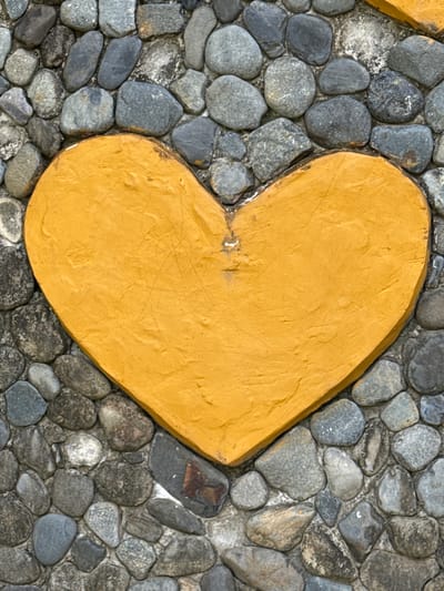 A close-up, overhead shot of a yellow, textured heart shape set into a wall of grey river stones. The heart is centrally located, taking up most of the frame. The stones are of varying sizes and shapes, with some smoother and rounder, while others are more angular and rough. The heart has a matte finish and appears to be made of painted concrete or stucco. The overall impression is one of rustic charm and simple decoration. The location is the city of Nam Khanh Vinh, Vietnam.