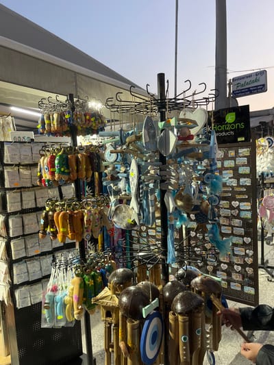 The image shows a souvenir stand in Limenaria, Greece (latitude: 40.626, longitude: 24.574). The stand is located on a street, with a building to the right and an awning overhead. It is filled with colorful souvenirs, including wooden fish, keychains, small wooden wind chimes, and small trinkets on display boards. A person is shown on the right side of the image, reaching toward the wind chimes. The stand is owned by "Greek Horizons Products." A sign for a restaurant "Restaurant - Grill Palataki" is visible on the building to the right.