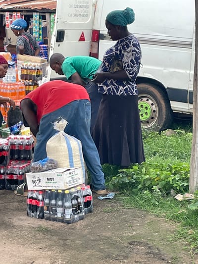 The image depicts an outdoor market scene in Wang, Nigeria. In the foreground, a man wearing a red shirt and blue jeans is bent over, seemingly arranging or inspecting goods. Beside him is a large white and blue striped bag, possibly containing rice or flour, and a box labeled "MOY LAUNDRY SOAP". Stacks of bottled beverages, including Coca-Cola and other dark-colored drinks, are arranged on the ground around the man. To the left, another person, partially visible, is wearing a red headscarf. In the background, a white van is parked, with some writing on its side that is partially obscured. Further to the left, a woman in a blue headscarf and patterned clothing is standing, looking towards the left. The ground is a mix of dirt and grass, with some vegetation growing. The overall impression is a busy, informal market setting.