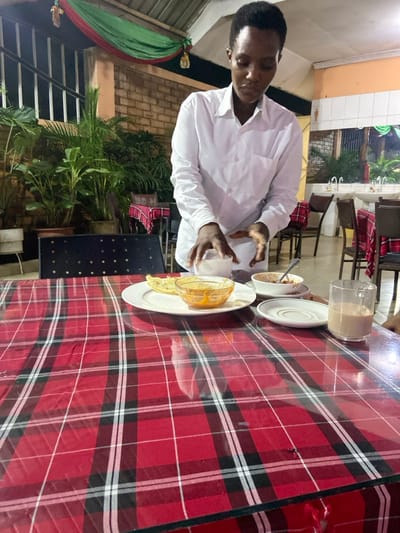 An individual is depicted seated at a dining table, likely enjoying a meal or snack in what appears to be an outdoor or semi-outdoor restaurant or cafe setting in Bagamoyo, Tanzania. The person, a man with dark skin and short dark hair, is wearing a white long-sleeved shirt. He is captured mid-action, holding a white object, possibly a piece of bread or a napkin, over a plate.
The table is covered with a distinctive red, black, and white plaid tablecloth. On the table, various food items are visible: a clear glass containing a light brown beverage, which could be tea or coffee with milk; a small white bowl holding a brownish, textured food, perhaps porridge or a stew; and a white plate presenting what appears to be pancakes or flatbread.
The background reveals a relaxed dining environment. Other tables and chairs, some with matching plaid seat covers, are faintly visible further back. Lush green potted plants and foliage contribute to a tropical ambiance. To the left side of the frame, a green and red striped hammock is partially seen. The lighting in the scene is artificial, suggesting the image was taken during the evening or at night. There is no visible text in the image.