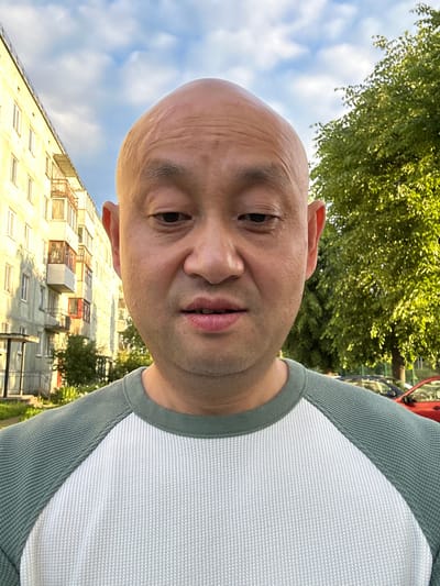 This is a selfie taken outdoors in Kirovsk, Russia. The image features a close-up of a bald man's head and shoulders, as he is looking directly at the camera. He appears to be in good spirits, with a slight smile on his face. The man is wearing a light-colored shirt with green trim. 

Behind the man, a residential building can be seen, with balconies and windows. Trees and a blue sky with clouds are also visible in the background, suggesting it is daytime and the weather is fair. The image is taken from a low angle, looking up at the man. There is no visible text in the image.