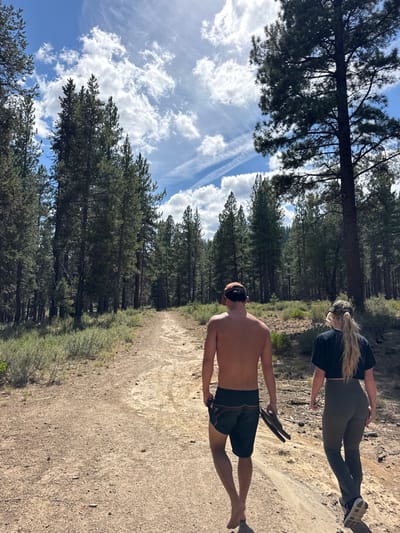 The image displays an outdoor scene on a dirt trail within a dense coniferous forest, consistent with environments found in or near Bend, Oregon, United States. Two individuals are depicted walking away from the camera, proceeding down the center of the unpaved path. The subject on the left is a male, shirtless, wearing dark shorts and a backward orange baseball cap; he is barefoot and holds a pair of dark sandals in his left hand. The subject on the right is a female, wearing a dark short-sleeved top, dark leggings, and a camouflage baseball cap; she is wearing dark athletic shoes. Both subjects are moving in the same direction, away from the viewer. The environment features numerous tall pine trees on either side of the trail, extending into the background. The ground is dry earth with scattered low-lying green and brown scrub brush. The sky above is partly cloudy with visible blue sections and several prominent contrails, indicating daytime conditions with direct sunlight.