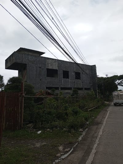 The image displays an outdoor scene in Melakudikadu, India, under an overcast sky. A multi-story concrete structure, appearing unfinished with exposed grey concrete walls, dominates the left-center. The building features multiple rectangular window openings without glass and a distinctive cantilevered section on its left side. In the foreground, a fence composed of vertical wooden planks and wire mesh separates a dense area of green vegetation and dirt from a paved asphalt road. Numerous black utility lines crisscross diagonally across the upper portion of the frame. On the far right side of the road, a light-colored pickup truck is partially visible, loaded with several large white sacks, and appears to be moving away from the viewer. Beyond the road and vehicle, trees and a utility tower are present in the background. No individuals are visible.