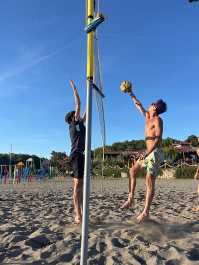 Two young men are playing beach volleyball on a sandy court in Pistula, Montenegro. The man on the left is wearing a dark t-shirt and black shorts, reaching up to block the ball. The man on the right, with purple hair, is jumping and spiking the ball over the net. He is shirtless and wearing patterned swim trunks. A volleyball net and poles are set up on the beach, with trees and buildings in the background. A playground with colorful slides is visible in the distance. The sky is clear and blue, indicating it is daytime.