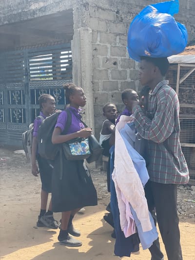 In Ikoga, Nigeria, a group of four people stand on a dirt-covered ground near a metal gate and a brick wall. Two females are in the foreground, dressed in purple shirts and dark skirts, each carrying a shoulder bag. Behind them are two males. One is dressed in a white tank top, and the other carries a stack of folded button-down shirts, with a large blue bag balanced on his head. The individual carrying the shirts wears a plaid jacket.