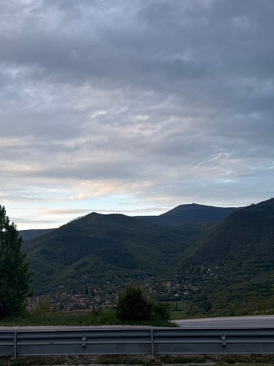 The image shows a landscape in Bunovo, Bulgaria, featuring a mountain range covered in dense green vegetation. A small village or settlement is nestled along the lower slopes of the mountain. A gray guardrail runs along the right side of the frame, indicating the presence of a road or highway. The sky is overcast with gray and white clouds. The photograph appears to have been taken at dusk or dawn, judging by the soft lighting.