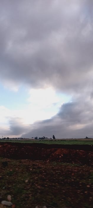 A rotated view of a landscape under a cloudy sky. Thick, grey clouds dominate the scene, with a bright, diffused light breaking through. Below the sky, a field with varying textures and colors extends. Parts of the field are green, while others show darker, reddish-brown tones, likely representing soil or tilled earth. The horizon line is indistinct.
