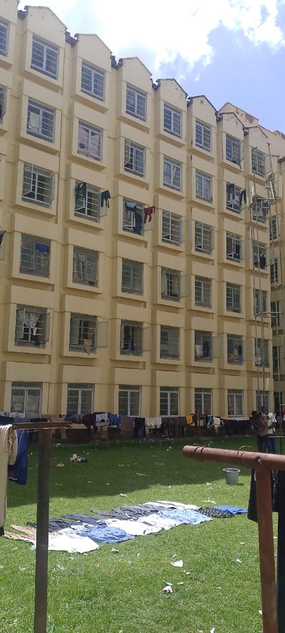 The image displays a daytime exterior view of a multi-story building and an adjacent grassy area in West Sakwa ward, Kenya. The building, painted in a light cream or yellow, consists of at least six visible levels, each featuring multiple identical windows secured with metal grates. Numerous windows are open, and various articles of clothing are observed drying from their sills and frames. The roofline is characterized by distinct peaked architectural elements, some exhibiting signs of wear or damage.
The foreground and midground consist of a green grass lawn, which is scattered with debris including paper and fabric scraps. Extensive laundry is present: numerous garments, predominantly light and dark-colored, are laid out in parallel rows on the grass to dry. Additional clothing hangs from a horizontal line positioned near the building's base and from a rustic, rusty metal drying rack located in the immediate foreground.
On the right side of the scene, two individuals are standing near the building's facade, adjacent to a tall, multi-level metal scaffolding structure. Their specific actions or interactions are not clearly discernible. A grey bucket rests on the grass nearby. The sky overhead is partly cloudy with patches of blue.