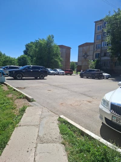 The image depicts a residential courtyard in the city of Jitiqara, Kazakhstan. In the foreground, a portion of a white car is visible, with its license plate reading "KZ 856 [illegible]". The car is parked on an asphalt surface, adjacent to a concrete curb overgrown with grass and weeds. In the midground, several cars are parked, including a black SUV, a red car, and various other sedans. Two individuals are walking in the distance. The background features apartment buildings, with some architectural details like a starburst pattern on one building. Trees and a clear blue sky complete the scene. The overall environment suggests a typical urban residential area under bright daylight conditions.