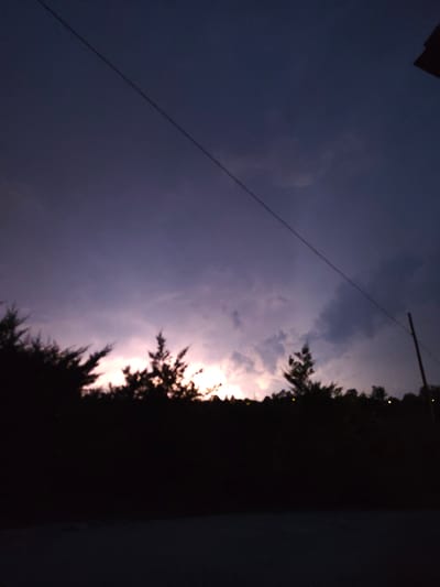 A diagonal power line stretches across a dark purple sky with some faint clouds. Silhouetted trees and bushes form a dark line across the lower third of the image. In the distance, the sky is illuminated by a pale pink and orange glow, suggesting a sunset or sunrise. A single utility pole is visible on the right side of the frame. The foreground is a dark, indistinct area, possibly a road or ground. There are no people or specific structures of Guarene, Italy, visible in the image.