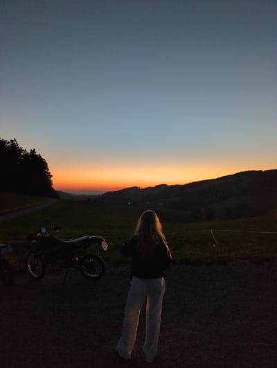 An image captured at dusk, likely in the vicinity of St. Peter, Germany, showcases a scenic overlook. A young person with long, blonde hair stands with their back to the camera, facing a vast landscape. The sky displays a gradient of colors, transitioning from a deep blue at the top to vibrant orange and yellow hues near the horizon, indicative of sunset.
In the foreground, a dark-colored motorcycle is parked, its silhouette clearly defined against the darkening terrain. The ground appears to be a mix of gravel and patchy grass. The middle ground features rolling hills covered in grass and trees, with a winding path visible on the left side. The background reveals more distant hills and a treeline, all bathed in the fading light of the day. The overall atmosphere is serene, suggesting a moment of quiet contemplation amidst nature.