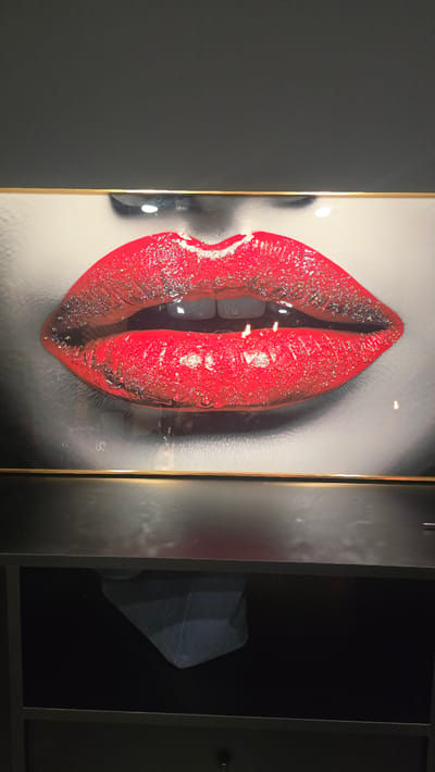 The image is a close-up of a framed artwork depicting bright red lips with glitter. The artwork is illuminated by overhead lighting, which casts reflections on the surface of the lips. The frame appears to be gold and black. Below the artwork, a dark shelf is visible, containing a dark blue fabric object. The context of the image suggests it is displayed in an indoor setting, possibly a home or a gallery, located in Trikomo, Northern Cyprus / Cyprus. There are no people or actions depicted.