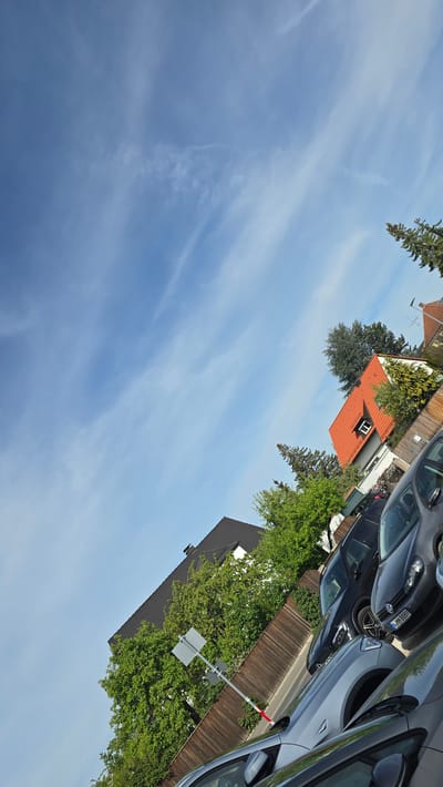 The image captures a residential street scene in Oberasbach, Germany, under a bright blue sky with faint cirrus clouds. Several cars are parked along the roadside, with license plates visible, including "N-K 8889." A wooden fence runs along the edge of the properties, and a gray metal pole supports a sign. Houses with red tile roofs and dark gray roofs are visible among trees and green foliage. The overall impression is a quiet, suburban neighborhood.