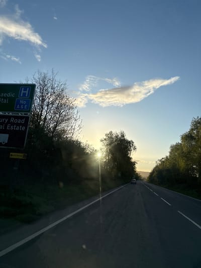 The image depicts a road in the United Kingdom, possibly near Oswestry, as indicated by the road sign pointing to "St. Oswald's". The road is straight and appears to be a dual carriageway. A single car is visible in the distance. The sun is low on the horizon, possibly during sunrise or sunset, partially obscured by trees and creating a lens flare. A prominent cloud formation is visible in the blue sky. Road signs for a "Paediatric Hospital" and "Maesbury Road Industrial Estate" are visible in the foreground, suggesting the location is near a hospital and an industrial area. The surrounding environment is characterized by trees and greenery.