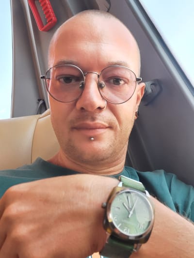 A man with a shaved head, wearing round metal-rimmed glasses, a septum piercing, and a lip piercing, is looking at the camera. He is wearing a dark green t-shirt and a watch with a light green face and a dark green strap on his left wrist. The man is seated in what appears to be the passenger seat of a vehicle, with a beige leather seat visible behind him and a grey interior panel. A red handle is mounted on the panel above and to the left of his head. The image is taken from a slightly low angle, suggesting the photographer is looking up at the subject. The lighting indicates it is daytime, possibly outdoors or near a window, with a light blue sky visible in the upper left corner. The man's hand is resting on his lap or armrest, with the watch prominently displayed. The image appears to be a selfie. There is no indication of the specific location within the city of Konya, Turkey.