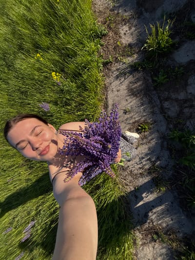 An overhead shot shows a woman lying in a grassy area, holding a bouquet of purple lupine flowers. She has light skin, closed eyes, and short dark hair. She is wearing a dark top and an earring is visible. Her arm extends out towards the camera. The location appears to be in a field near a concrete or paved area with some weeds growing in cracks. The grass is green, with scattered yellow flowers. Part of a dress or blanket with a black and white pattern and a white shoe are visible. The presence of lupines and the general vegetation suggest that this location could be in a rural or suburban area in Vakhrino, Russia.