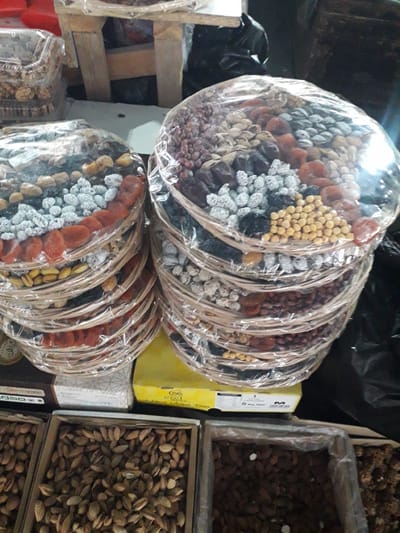 This media file captures a vibrant display of dried fruits and nuts, likely in a market stall in Ghafurov, Tajikistan. The foreground is dominated by two stacks of circular, shallow baskets, each meticulously arranged with various dried goods and individually wrapped in clear plastic for sale.

The top stack shows baskets filled with an assortment that includes light-colored dried mulberries, golden chickpeas, reddish-brown dried apricots, and darker dried fruits, possibly dates or black mulberries. Some sections also contain what appear to be red peanuts or another type of legume. The second, lower stack of baskets features a similar colorful variety, with prominent sections of white-dusted dried mulberries, vibrant orange dried apricots, and bright green pistachios, alongside other nuts and dried fruits.

To the right of the stacked baskets, several open wooden boxes are filled with bulk nuts and seeds. The top visible box contains numerous unhulled almonds, while the box below it seems to hold another variety of nuts or seeds, possibly unhulled pistachios or a similar item.

The background is somewhat cluttered, suggesting a commercial setting. A yellow cardboard box is visible between the two main displays, with partial text showing "5 kg Net" and the letter "M" on its side. Other items, such as black plastic bags and wooden structural elements, are dimly visible. The overall impression is that of a bustling market, with goods attractively prepared and presented for customers. The lighting appears to be artificial, casting reflections off the plastic wrapping. No people or specific activities beyond the display of goods are discernible.