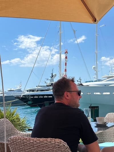 A man wearing sunglasses and a black t-shirt sits at an outdoor cafe table, facing away from the camera and looking out at a harbor. He is seated in a woven chair, and on the table in front of him is a glass with a straw and a cup of coffee. Behind the man, several large yachts are docked in the bright blue water under a clear sky with scattered clouds. A few seagulls are visible in the sky above the yachts. The scene is framed by a tan umbrella that casts a shadow over the top portion of the image. This image was taken in Donja Lastva, Montenegro.