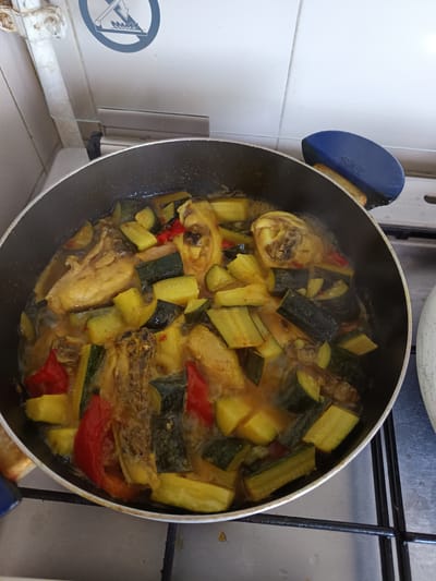 An overhead shot shows a black frying pan filled with chicken and vegetables. The chicken pieces are light brown and appear to be cooked, some showing bone. The vegetables include chopped zucchini, cut into thick half-moon shapes with dark green skin and lighter green flesh, and chunks of red bell pepper. The mixture is in a sauce that is a light golden brown. The pan is on a stovetop, with the burner grates visible below. The background shows a white tiled wall with a pipe and a circular warning sticker. The sticker has a blue border and a white center with a black symbol of a flame under a pan and a diagonal line through it, indicating "no open flame" or "no cooking." A blue pan handle rest is positioned over the edge of the pan. The scene is likely a kitchen in Roseto degli Abruzzi, Italy, based on the information provided.