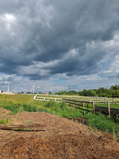 The image depicts a rural landscape under a heavily clouded sky, likely on the outskirts of Косяково, Russia. In the foreground, a large pile of dark brown organic material, possibly manure or compost, lies on the ground. A weathered wooden beam rests horizontally across this pile. Beyond the compost, a green field stretches out, interspersed with patches of tall grass and wildflowers. A wooden fence with vertical posts and horizontal rails runs through the middle ground, separating the field from a further area. To the left of the wooden fence, a white railing fence is visible. In the background, a dense line of green trees forms a treeline. Several power poles with wires are scattered across the horizon, and various buildings, including what appear to be houses with red roofs, are visible among the trees. The sky is dominated by dark, heavy clouds, with some lighter patches of blue sky and white clouds visible towards the horizon. The overall impression is one of a somewhat overcast day in a rural setting.