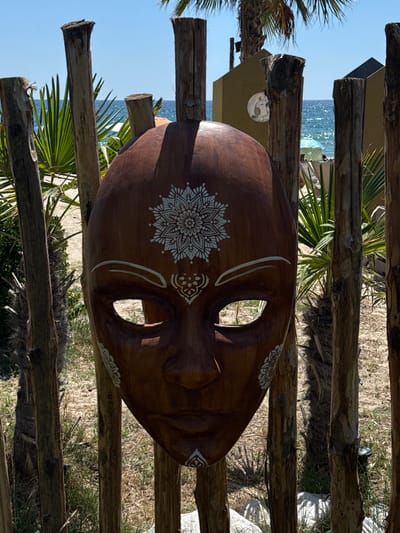 A wooden mask with white mandala-like designs is affixed to a rustic fence made of vertical wooden posts. The mask is dark brown and features cut-out eye openings and carved facial features. In the background, the blue sea meets a clear sky. Palm trees and sandy ground are visible, suggesting a beachside or coastal location. A building with a circular design on its wall is also present in the background. The overall scene appears to be outdoors on a sunny day, likely in Asprovalta, Greece.
