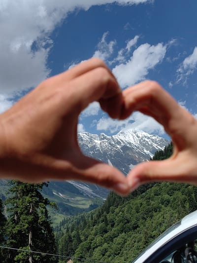 In Burwa, India, a pair of hands are positioned in the shape of a heart, obscuring the lower portion of a majestic snow-capped mountain range. The mountains are rugged, with visible rock faces and patches of snow, set against a vibrant blue sky dotted with white clouds. Below the peaks, a lush green forest covers the rolling hills, creating a dramatic contrast between the starkness of the mountains and the vitality of the vegetation. The lower edge of the frame shows a portion of a white vehicle, suggesting a scenic drive or viewpoint.