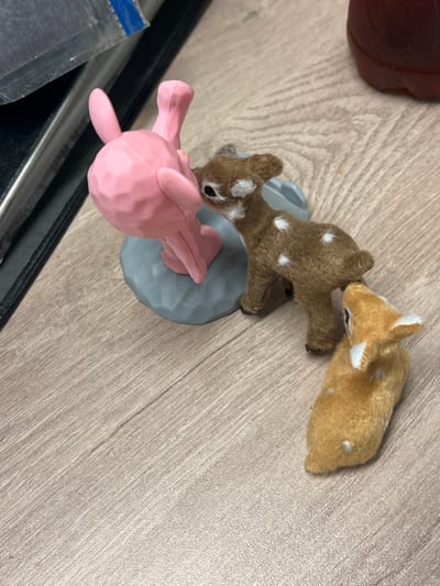 On a light brown, wood-patterned surface are three animal figurines. Two appear to be fuzzy toy deer, one brown with white spots and one tan. These deer figurines are posed close together. Next to them is a stylized, geometric-looking pink elephant figurine. The deer and elephant figurines are arranged on a grey base. Part of a brown object is visible in the upper left and part of a silver object in the lower left of the scene.