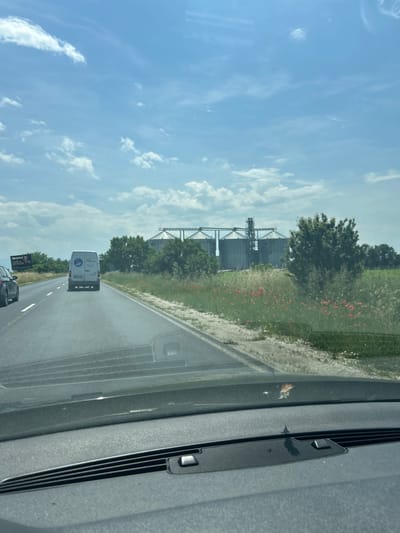 The image, taken from inside a moving vehicle, captures a daytime scene along a two-lane asphalt road, likely in Graf Ignatievo, Bulgaria. The sky is bright blue with scattered white cumulus clouds.
In the foreground, part of the car's interior, specifically the passenger-side dashboard and windshield frame with a wiper assembly, is visible. A small insect or piece of debris is present on the windshield.
On the road ahead, a white delivery van with a logo on its rear is visible. The logo depicts a stylized figure holding a package, with the word "RAPIDO" potentially visible below it, followed by smaller text. Behind the van, a dark-colored car is also traveling.
To the left of the road, a vibrant green landscape unfolds, featuring numerous trees and patches of bright red flowers, possibly poppies. Further in the distance, large, dark industrial silos, likely for grain storage, stand prominently against the sky, supported by intricate metal structures.
To the right of the road, a billboard is partially visible, clearly displaying the text "METRO" and "HIPERMARKET", indicating the presence of a supermarket nearby. The overall scene suggests a journey through a mixed agricultural and industrial area.