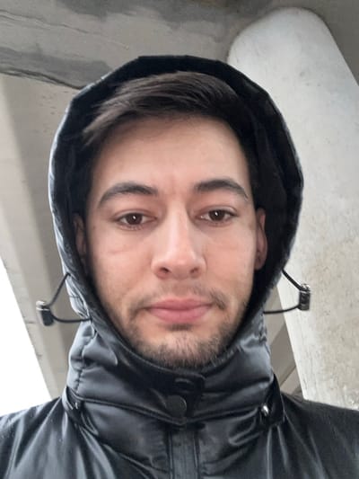 The image displays a close-up, head-and-shoulders portrait of a male individual standing outdoors in Yangiiul, Uzbekistan. The man, with light stubble and short dark hair visible beneath a raised hood, looks directly forward with a neutral expression. He is wearing a dark, possibly black, hooded puffer jacket with a high collar and visible snap buttons. Drawstrings with toggles are present on either side of the hood. The background consists of out-of-focus concrete structures: a flat, dark concrete underside of a large structure to the upper left and a lighter, thick concrete pillar or support to the upper right. The scene is illuminated by ambient light, suggesting an overcast environment or shade. No specific actions or interactions beyond the subject posing for the photograph are visible.