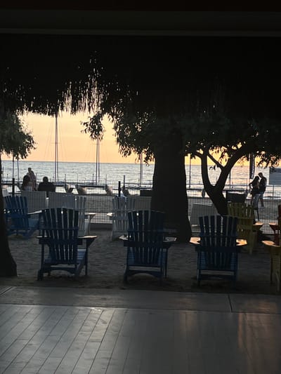 The image depicts a waterfront scene at sunset in Selçuk, Turkey, viewed from under a dark, shaded overhang. The foreground consists of a dark, horizontally planked floor, transitioning to a sandy area adorned with numerous Adirondack chairs of various colors, predominantly blue and white, arranged in pairs with small tables. Dark, silhouetted trees with visible leaf structures frame the central and right portions of the midground. A chain-link fence separates the sandy area from a body of water, which reflects the warm orange and yellow hues of the setting sun. Silhouettes of sailboats or yachts with prominent masts are visible on the water against the bright sky. Several individuals are present: a group of three to four people are observed near the fence on the left, and a pair of individuals stand closely together near the water's edge on the right, partially obscured by a tree. The overall lighting is back-lit, rendering most objects and people in silhouette against the vibrant sunset backdrop.